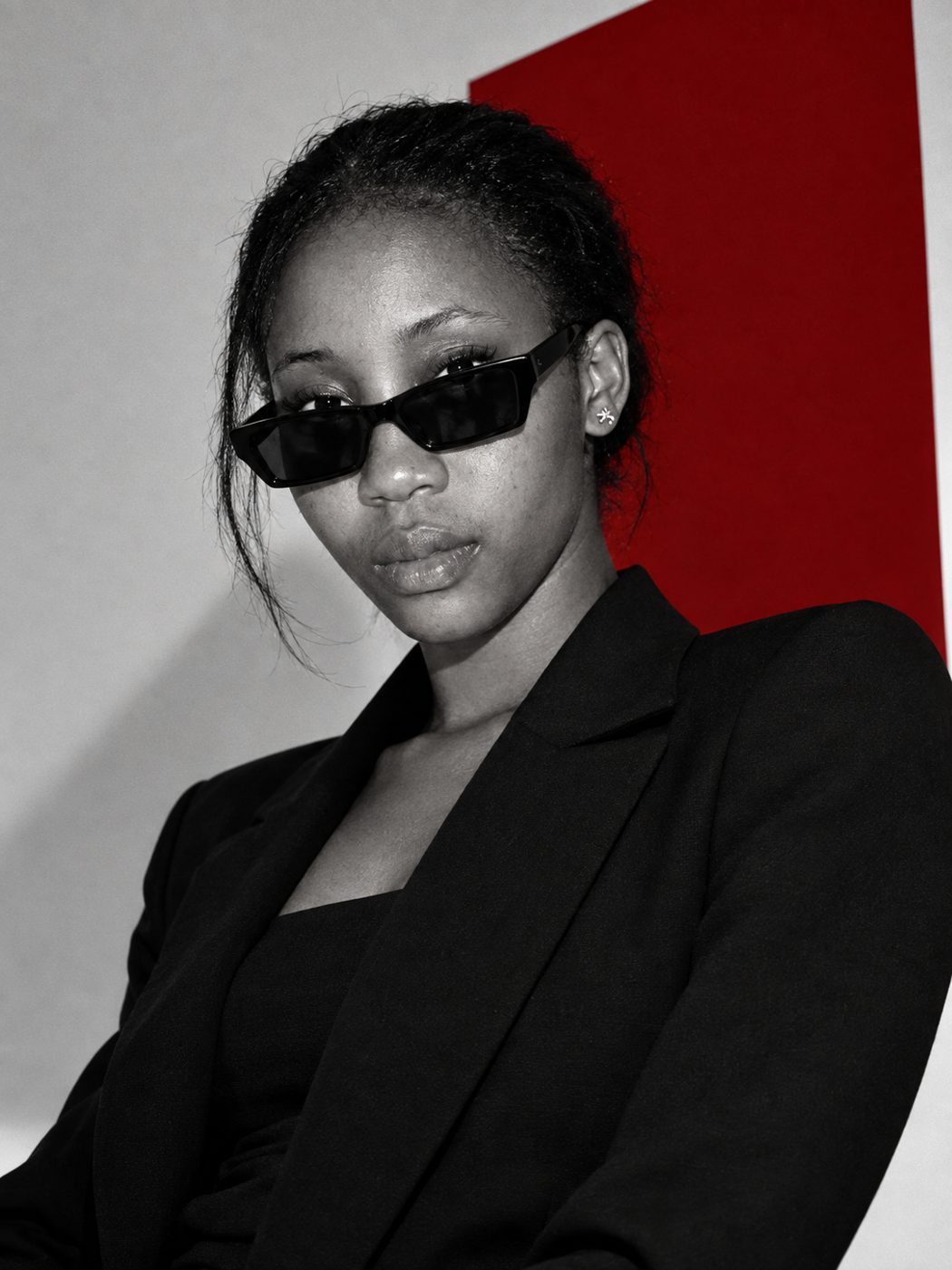 Black and white portrait of a woman in sunglasses and a black blazer against a maroon panel